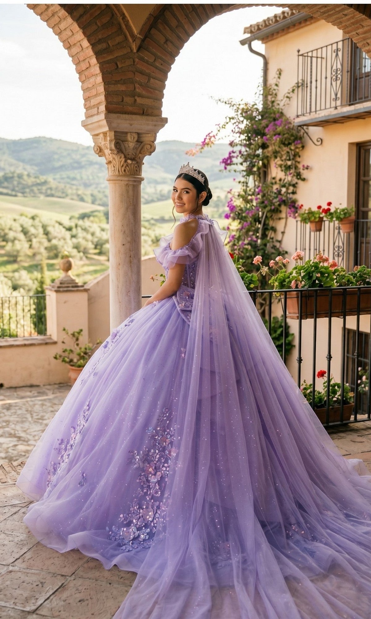 A woman wearing the Quinceanera Dress 8585JQ by Cinderella Couture, featuring a lavender ball gown with a glitter tulle skirt and a delicate cape, stands on a terrace adorned with flowers and scenic hills in the background, her look completed by a sparkling tiara.